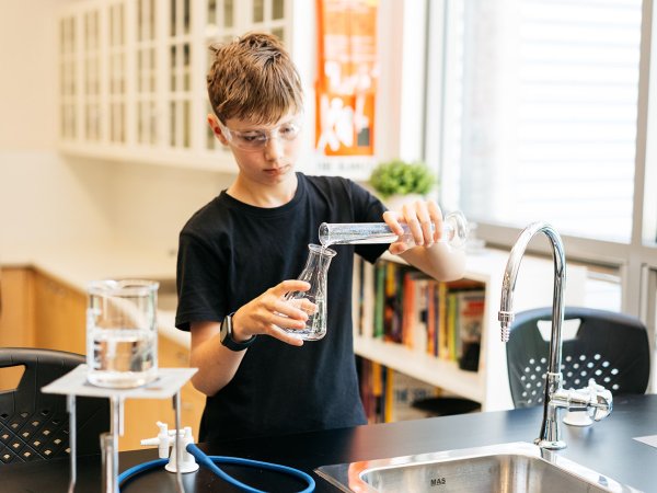 Child in a science classroom pouring liquid into a jug with safety glasses on Image